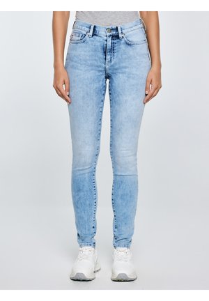 Asmuo dėvi šviesiai mėlynas siauras džinsus ir baltus sportbačius, stovi priešais šviesų, vienspalvį foną, rankos atsipalaidavusios šalia kūno.