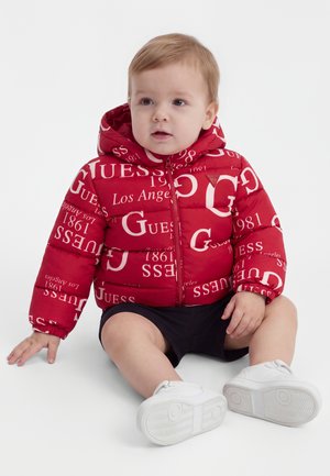 Малко дете, седнало, облечено с червено яке Guess с бял надпис, черни къси панталони и бели обувки пред светъл едноцветен фон.