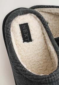 Zapatillas grises de felpa con un suave forro de crema, forma redondeada y ribete negro. Presentan una etiqueta negra con el nombre de la marca "NEXT."