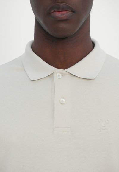 T-shirt polo beige clair en tissu texturé, col court et trois boutons. Présente un petit logo brodé sur la poitrine.