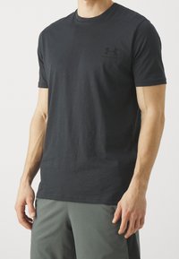 Muž v černém tričku s krátkým rukávem Under Armour a šedých sportovních šortkách stojí proti jednobarevnému pozadí.