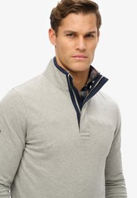 Grijze pullover trui met een ritssluiting bij de kraag, voorzien van een marineblauw accent en een geruit patroon eronder. Zachte stoftextuur en een casual pasvorm.