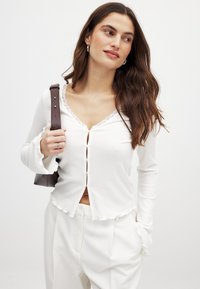 Weißes, geripptes Langarmoberteil mit einem V-Ausschnitt mit Spitze, Knopfverschluss und Rüschensäumen. Kombiniert mit weißen Hosen und einer braunen Handtasche.
