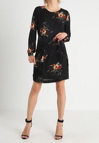 Vestido negro con mangas largas, patrones florales en rojo y amarillo, detalle de nudo en la cintura y una textura suave. Combinado con tacones negros.