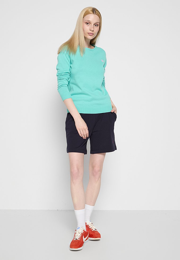 Junge Frau steht mit den Händen in den Taschen, trägt einen türkisfarbenen Pullover, schwarze Shorts, weiße Socken und rote Turnschuhe vor einem weißen Hintergrund.