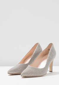 Srebrne szpilki z teksturowanej metalicznej tkaniny, zakończone szpiczastym nosem i eleganckim obcasem w stylu stiletto. Wnętrze jest miękkie i beżowe.