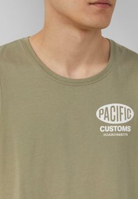 Olivgrünes Baumwoll-T-Shirt mit Rundhalsausschnitt, das auf der Vorderseite ein weißes bedrucktes Logo mit der Aufschrift "PACIFIC CUSTOMS BOARDS&KITS" zeigt.