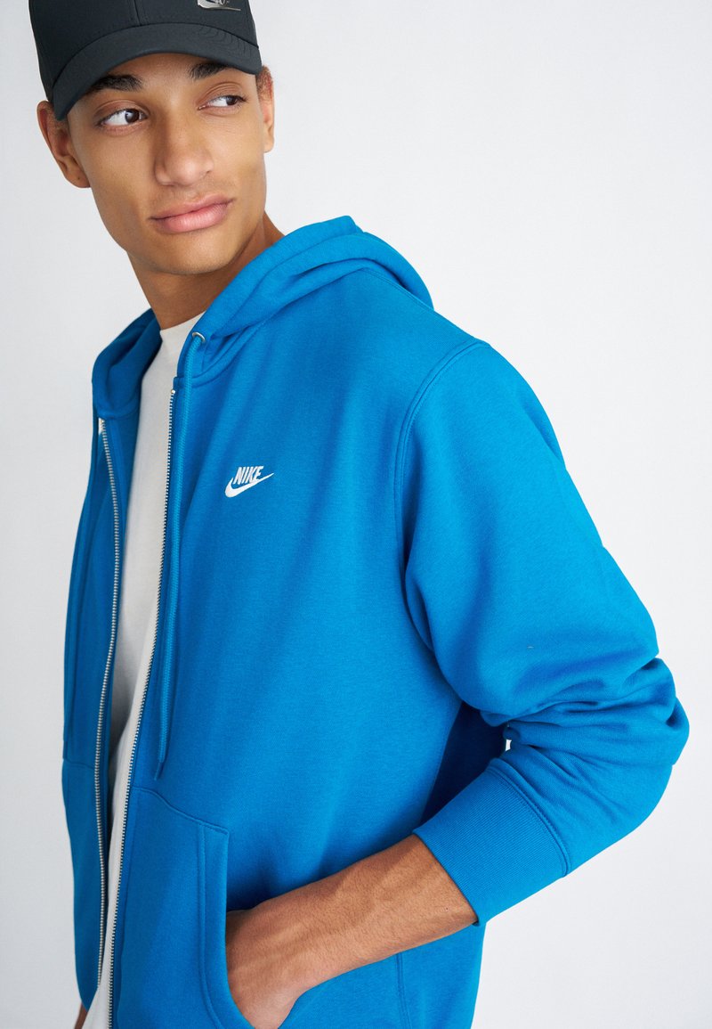 Sudadera con cremallera azul, hecha de un material suave, con un bolsillo delantero y capucha con cordón. Presenta un logo de Nike blanco en el pecho.