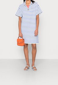 Pasek polo w kolorze białym i niebieskim z krótkim rękawem, w połączeniu z małą pomarańczową torebką i niebieskimi sandałami. Gładka struktura tkaniny.