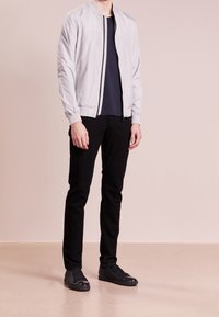 Chaqueta bomber gris claro con cremallera frontal, puños acanalados, usada sobre una camisa azul marino, combinada con vaqueros negros y zapatillas negras con textura.