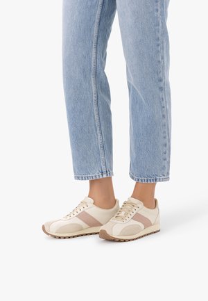 Person wearing light blue jeans and cream sneakers with beige accents standing on a white background.