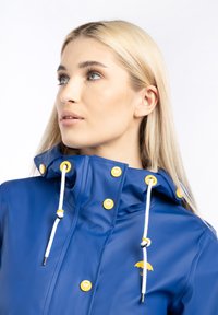 Femme blonde portant un imperméable bleu avec des boutons jaunes et des cordons blancs, regardant sur le côté devant un fond blanc.