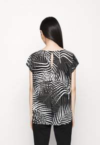 Donna con lunghi capelli scuri che indossa una blusa a maniche corte con stampa tropicale bianca e nera vista di spalle su uno sfondo bianco.