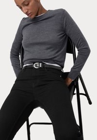 Graues langärmliges Strickoberteil, kombiniert mit hochgeschnittenen schwarzen Jeans und einem schwarzen Gürtel mit einer silbernen Schnalle, sitzt auf einem schwarzen Stuhl.