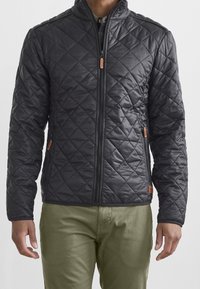 Veste noire matelassée avec un col haut, une fermeture éclair frontale et deux poches latérales ; comporte des accents en cuir marron et une texture lisse et brillante.