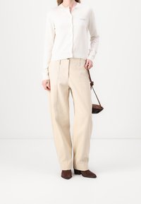 Bílý pletený cardigan s kulatým výstřihem, doplněný béžovými kalhotami s širokými nohavicemi. Doplňkem je malá hnědá taška přes rameno. Hnědé boty na podpatku.