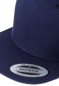 Mørkeblå snapback-cap laget av strukturert stoff med flat skygge, med en sirkulær metalllogo-patch og broderte detaljer.