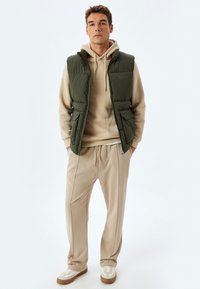 Grøn puffervest over en beige hoodie, parret med beige bukser med snøre og hvide sneakers med creme detaljer. Enkel, moderne design.