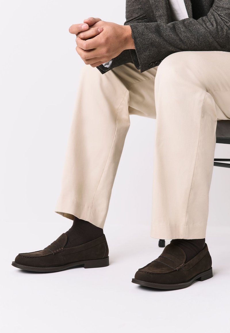 Homme assis, mains jointes, portant un pantalon beige, des mocassins marron foncé et un blazer texturé foncé.
