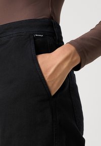 Črne denim hlače s prilegajočim pasom, podrobnostmi sprednjih žepov in majhno oznako blagovne znamke, ki je vidna na pasu. Gladka tekstura.