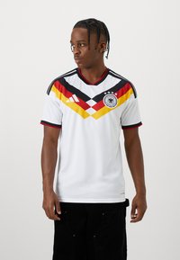 Homme portant un maillot de football allemand blanc avec un design en chevrons noir, rouge et jaune, debout devant un fond clair uni, regardant vers la gauche.