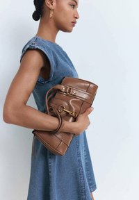 Geantă de piele maro cu detalii din metal auriu, ținută lângă o rochie din denim cu un croi structurat și mâneci rulote.