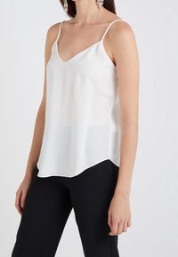 Femme portant un top en satin blanc sans manches avec de fines bretelles et un pantalon noir, se tenant contre un fond uni clair.