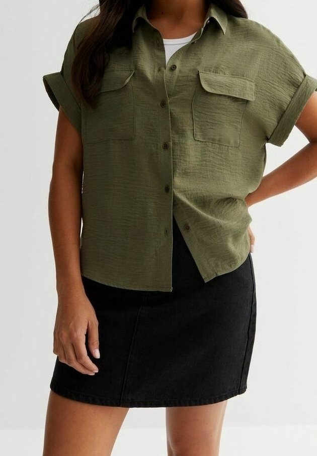 Femme portant une chemise boutonnée à manches courtes couleur vert olive avec des poches poitrine et une mini-jupe noire, debout devant un fond uni.
