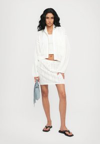 White knitted jacket with a cropped design, paired with a matching white skirt. Model holds a light blue bag, styled with black flip-flops.