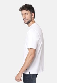 Camiseta blanca de algodón con mangas cortas, cuello redondo, aberturas laterales en el dobladillo y un corte relajado, combinada con jeans de mezclilla oscura.