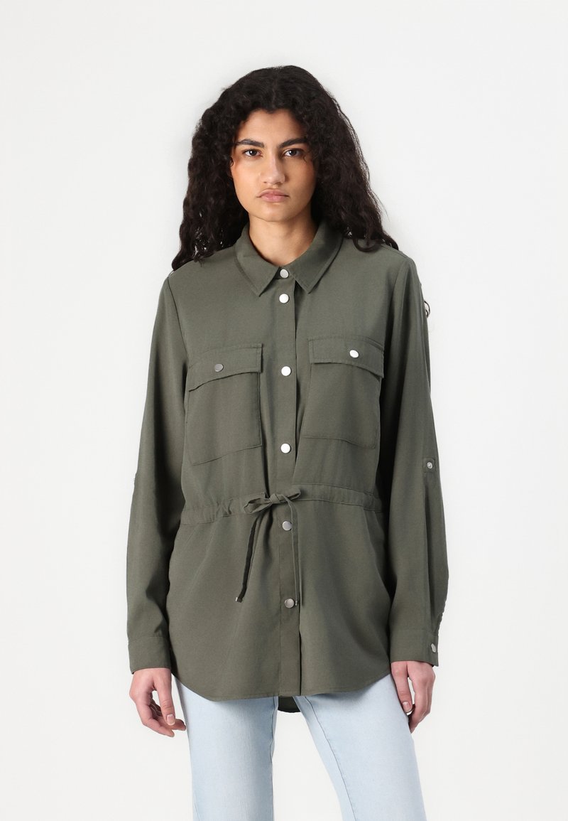 Olijfgroene, langärmige blouse met een knoopsluiting aan de voorkant, twee borstzakken en een verstelbare trekkoord taille. Gladde stof met zilverkleurige hardwaredetails.