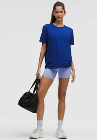 Blaues Baumwoll-T-Shirt kombiniert mit helllila Sportshorts, hält eine schwarze Sporttasche. Weiße Sneakers und graue Socken runden das Outfit ab.