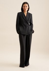 Czarna marynarka dwurzędowa z dwiema przednimi kieszeniami, połączona z szerokimi spodniami. Gładka tekstura, dopasowany krój, minimalistyczny design.