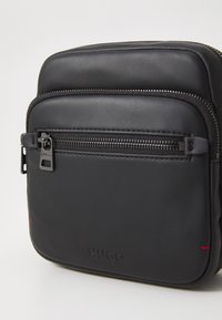 Borsa a tracolla sintetica nera con finitura liscia, tasca frontale con zip e branding sottile con cuciture di accento rosse.