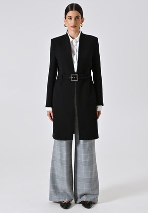 Cappotto lungo nero con vita cinturata, abbinato a pantaloni a gamba larga in plaid grigio e bianco, completato da scarpe nere.
