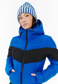Modrá a čierna izolovaná bunda s kapucňou, so zapínaním na zips, diagonálnym farebným blokovaním a dvoma bočnými vreckami. V páre so pruhovanou pletenou čiapkou.