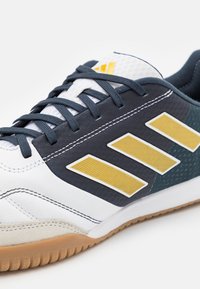 Zapatilla deportiva blanca con suela azul marino y de goma, que presenta tres rayas amarillas de acento, parte superior de sintético texturizado y cordones blancos.