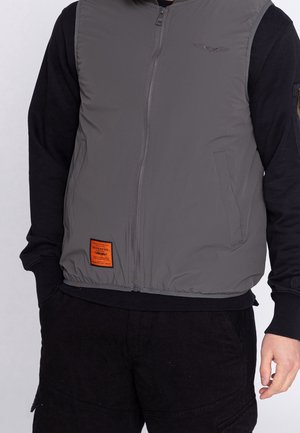 Mand iført en grå vest med lynlås og forlommer over en sort langærmet skjorte og sorte bukser, med hænderne i lommerne.