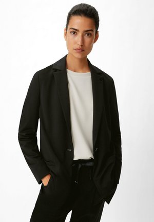 Schwarzer Blazer mit strukturiertem Design, Reverskragen und silbernen Hardware-Akzenten, kombiniert mit einem hellgrauen Oberteil aus weichem Material.
