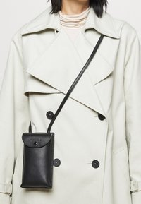 Osoba oblečená v krémovém trenčkotě a béžovém roláku, s malou černou koženou crossbody taškou na přední straně.