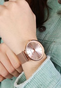 Rosé-goldfarbene Uhr mit einem Netzarmband, ausgestattet mit einem reflektierenden Rosé-goldenen Zifferblatt, schlanken Stundenmarkierungen und einem minimalistischen Design. Getragen an einem Handgelenk mit einer mintfarbene Cordjacke.