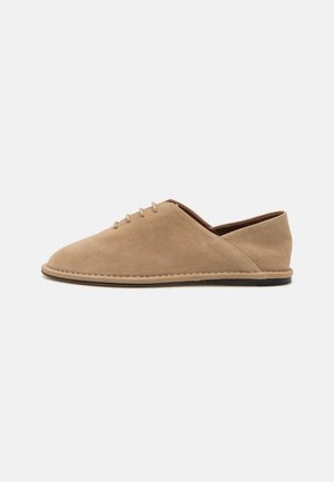 Beige semskede sko med lav profil, rund tå og en enkel snøring foran. Siden har en liten utskjæring.