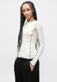 Biały top z długim rękawem z czarnymi, falistymi krawędziami, wykonany z miękkiego materiału. Posiada okrągły dekolt i dopasowaną sylwetkę. W zestawie z czarnymi spodniami.