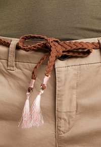 Flätad brun mockabälte med rosa fransar som är fäst i midjan, som kompletterar ljusbeige byxor och en olivgrön topp. Texturen synlig.