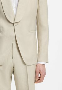 Beżowa, dopasowana marynarka na jeden guzik, do kompletu spodnie oraz biała mankiet koszuli widoczna na ramieniu i ręce osoby.