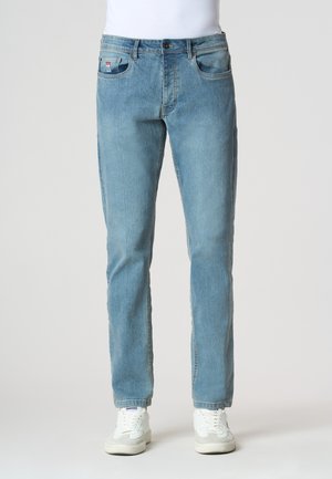 Jeans azzurri a gamba dritta indossati con sneakers bianche e una maglietta bianca, mostrati frontalmente su uno sfondo semplice.