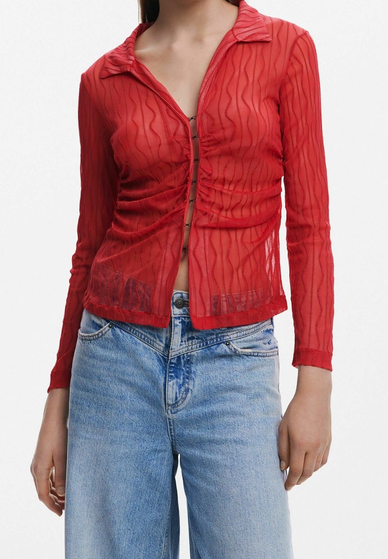 Femme portant un haut transparent rouge à manches longues avec des motifs ondulés, associé à un jean taille haute bleu clair.