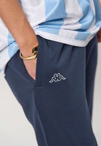 Osoba w niebiesko-białej koszuli w paski, z ręką w kieszeni granatowych spodni, pokazująca logo marki Kappa i złotą bransoletę na nadgarstku.