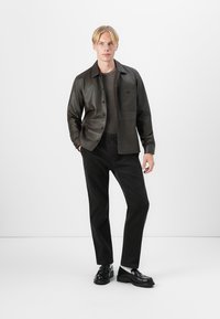 Dunkles Lederhemd mit Knöpfen und einer Brusttasche, getragen über einem eng anliegenden grauen Oberteil; kombiniert mit schmal geschnittenen schwarzen Hosen und schwarzen Schuhen.