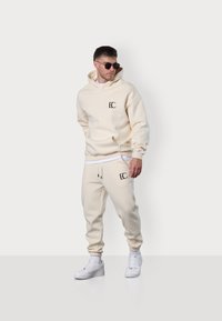 Beige Kapuzenpullover und Jogginghose aus weichem Material, mit einer frontalen Tasche und Logo; das Model trägt weiße Sneakers und Sonnenbrille.